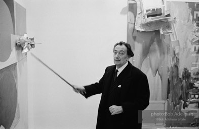 Salvadore Dali at a Jasper Johns exhibit at the Leo Castelli gallery using his cane to point to an element in John's assemblage. Circa 1966. New York City.